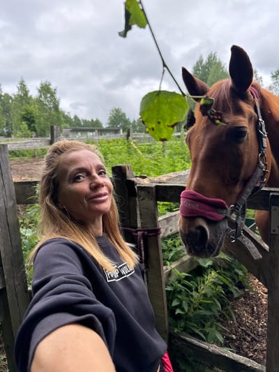 In Лемболово-парк Днп, Russia, a woman with blonde hair, wearing a black sweatshirt, stands next to a brown horse. The woman is holding a phone and taking a selfie, with the horse's head visible on the right side of the frame. The horse is wearing a pink noseband and a halter. They are in an outdoor setting, likely a stable or farm, with a wooden fence and lush green vegetation in the background. The sky is overcast, suggesting a cloudy day.