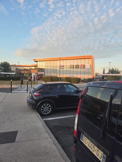 In Briis-sous-Forges, France, a dark-colored Nissan Juke is parked on the left side of the frame, facing away from the viewer. To the right, a dark blue Mercedes-Benz van is parked, its rear visible. The van has a license plate that reads "DC-445-GJ 02". In the background, a modern building with a glass facade reflects the sky. Several businesses are visible on the building's exterior, including "Bonjour," "McDonald's," "Paul," "Pizza Hut Express," and "O'Table!". People are seen entering and exiting the building. The environment suggests a roadside service area or a commercial complex. The sky is a clear blue with scattered clouds, indicating either early morning or late afternoon. The lighting is soft, suggesting a sunset or sunrise.