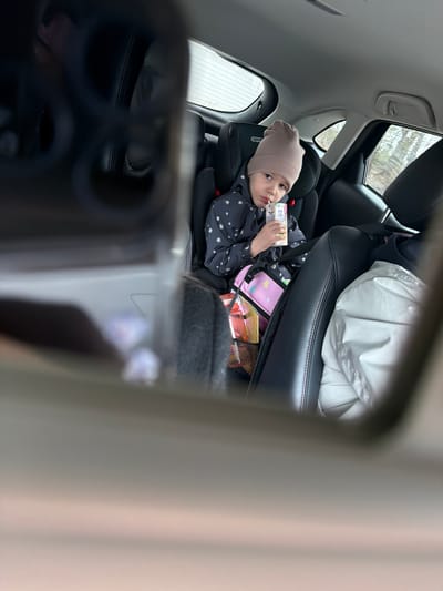 A young child wearing a beige hat is in a car seat, drinking from a juice box. The car seat is in the back of a vehicle, behind a larger seat covered with a white jacket. A pink backpack and other items are visible near the child's car seat. The image appears to be taken from the front seat through a car mirror. No specific location details relating to the city of Kukui, Russia are discernable from the image.