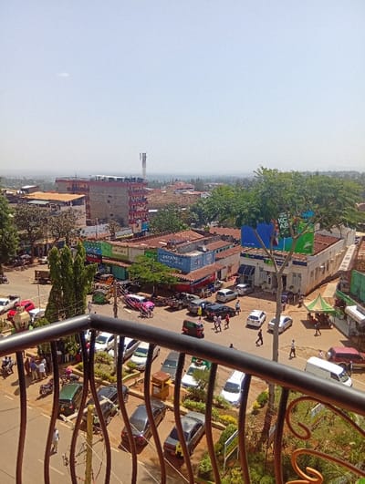 This elevated daytime image captures a busy urban scene in Murang'a, Kenya, viewed from behind an ornate, dark metal railing.

The foreground is occupied by a dark, decorative metal railing with vertical bars and scrollwork, partially obscuring the lower street level.

The midground displays a network of asphalt roads with numerous vehicles and pedestrians. Parked vehicles include approximately two dozen cars of various makes and colors (white, silver, black, red, green), a white delivery van, a pink motorcycle, and a green auto-rickshaw. Pedestrians, numbering over twenty, are observed walking on sidewalks and crossing roads. Commercial buildings line the streets, featuring visible signage such as "PAYANI ENTERPRISES" on a blue container-like structure and a partial "KCB BANK" sign on a white building. A green tent structure is erected roadside on the right. Trees with green foliage and landscaped areas with shrubs are interspersed throughout the street level. Utility poles with wires are also visible.

The background reveals a densely packed urban area with multi-story buildings, including a prominent red and white apartment block. Rooftops feature water tanks. A tall, lattice-style communication tower stands against the clear, light blue sky on the distant horizon, which is marked by hazy landforms. The overall scene is brightly illuminated by direct sunlight, casting short shadows.