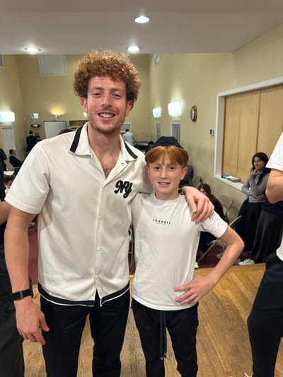 This indoor image captures an adult male and a child male posing in what appears to be a community hall or synagogue in Bergenfield, United States. The adult male, positioned on the left, has curly reddish-brown hair and a light beard, wearing a white short-sleeved collared shirt with black trim and a black "NY" emblem on the left chest, over black pants. He also wears a black wristband on his left arm and smiles directly at the camera with his right arm around the child's shoulders. The child male, positioned on the right, has red hair with bangs and wears a black kippah, a white short-sleeved t-shirt with "SONNETTI LONDON" printed on the chest, and black pants with a grey drawstring. He also smiles at the camera with his left hand on his hip. The background features light-colored walls, overhead lighting, wall sconces, and a polished wooden floor. Several other individuals, mostly blurred, are visible in the background; some are seated at tables or standing, and a woman in a grey top and dark skirt is visible near a window on the right. A clock is visible on the far right wall. A projector screen is partially visible on the far left.