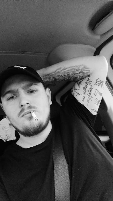 This black and white photograph, taken in Senec, Slovakia, depicts a male subject seated within what appears to be the interior of a vehicle. The male is positioned centrally, looking directly at the camera. He wears a dark baseball cap with a visible light-colored logo and a dark t-shirt. Facial hair, including a beard and mustache, is present. A lit cigarette is held between his lips. His right arm is bent and raised, with his hand possibly resting behind his head or against the vehicle's interior. Extensive tattoos are visible on his right arm, including a full sleeve pattern on the upper arm and script reading "There is one authority for me - my father!" on the inner bicep area, alongside other designs. A dark seatbelt is taut across his chest. The vehicle's headliner is visible in the upper background, and a vehicle pillar or door frame is partially visible on the right. The overall scene is a close-up, likely a selfie, taken in a low-light or shaded environment.