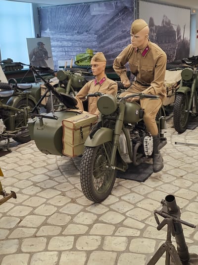 The image depicts an exhibit in Архангельское, Russia, featuring vintage military motorcycles with sidecars. Two mannequins dressed in period military uniforms are positioned on the motorcycles, simulating riders. The motorcycles are olive green and appear to be from the World War II era or shortly after. In the background is a large mural depicting soldiers in battle. The floor is a light-colored stone pattern. A mortar is situated on the right side of the image.