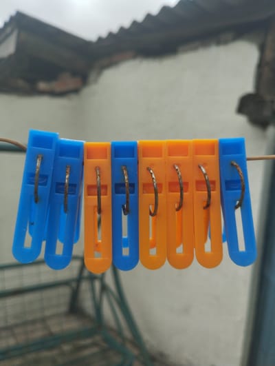 A close-up, eye-level shot captures a string of seven plastic clothespins, alternating between blue and orange, attached to a thin wire. The clothespins are positioned horizontally across the frame, with their open ends facing downwards. Each clothespin features a rusted metal spring mechanism. In the blurred background, a rough-textured, light-colored wall is visible beneath a dark, corrugated roof structure. The scene appears to be outdoors, possibly in a residential area of Lebedyn, Ukraine, given the context.