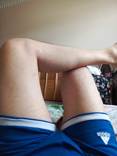The image shows a close-up of a person's legs, which are bare and lightly covered in hair. The person is wearing blue athletic shorts with a white waistband and the Adidas logo visible on the right leg. The legs are crossed, with the left leg resting on top of the right. The background is blurred but suggests an indoor setting. Behind the legs, there is a wooden dresser with decorative carvings, a bed with patterned bedding, and a glimpse of a black garment with a floral print. The lighting appears to be natural, coming from a window or a well-lit room, suggesting it could be daytime. The overall impression is casual and relaxed.