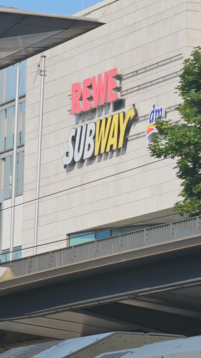 The image shows a modern building in the city of Neu-Ulm, Germany. The building features a light-colored, stone-tiled facade with horizontal lines. Prominently displayed on the facade are large, red, three-dimensional letters spelling "REWE," a German supermarket chain. Below "REWE," also in three-dimensional letters, is the word "SUBWAY" in white with a yellow arrow pointing to the right. To the right of "SUBWAY" and slightly above it, the logo for "dm," a drugstore chain, is visible in blue. A green tree with dense foliage extends into the right side of the frame, partially obscuring the upper right portion of the building. In the foreground, a metal walkway or bridge with a railing and vertical supports spans across the lower part of the image, casting a shadow on a structure below. The sky is clear and blue.