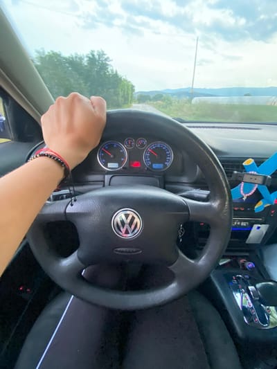 The image shows a first-person perspective from inside a car, with the driver's hand on the steering wheel. The steering wheel is black with a silver Volkswagen logo in the center. The dashboard displays a speedometer reading around 70 km/h and a fuel gauge. To the right of the steering wheel, a blue air freshener in the shape of a cross is visible, adorned with a pink necklace. The car is driving on a road with trees and fields on either side, under a cloudy sky. The scene does not provide any specific details to indicate the location in the city of Miriantsi, Bulgaria.