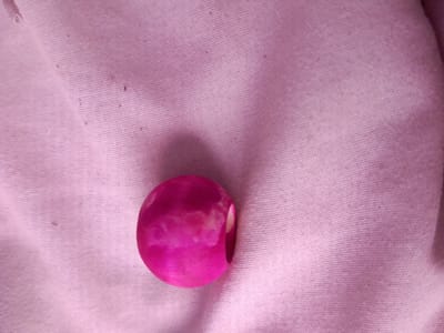 The image displays a close-up, overhead view of a single, smooth, oval-shaped object with a vibrant magenta hue. The object rests on a textured, pale pink fabric that appears to be cotton or linen, exhibiting a subtle ridged pattern. The lighting casts a soft shadow to the left of the object, highlighting its rounded form. The fabric itself is slightly creased in places, indicating it is likely a piece of clothing or bedding. There are a few small specks of dark debris scattered on the fabric, suggesting the surface may not be entirely clean. The object's surface has a slight sheen and some lighter, irregular white markings are visible within the magenta color, possibly indicating marbling or a translucent quality. There is no indication of the city of Inhulo-Kamianka, Ukraine, in this image.