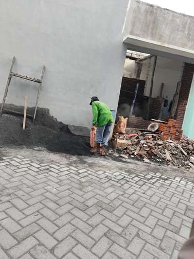 In Katrungan, Indonesia, a construction worker wearing a green long-sleeved shirt and blue pants is shown near a pile of dark granular material. The worker is barefoot and appears to be handling a bag of what looks like cement. In the background, a wall of a building under construction is visible, with some exposed brickwork and debris. A wooden structure, possibly a sieve, stands against the wall next to the pile of material. The ground in the foreground is paved with interlocking concrete blocks arranged in a herringbone pattern. The scene depicts an ongoing construction project in an outdoor urban environment.