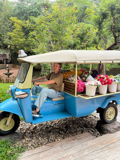 In Mae Rim, Thailand, a man sits in the driver's seat of a blue and beige tuk-tuk that is filled with a colorful assortment of flowers in buckets and wooden crates. The tuk-tuk is parked on a bed of decorative gravel and appears to be in a garden setting, with lush green trees forming the backdrop. The man is wearing a light olive green t-shirt and light blue jeans. The tuk-tuk has a cream-colored canopy and a white sign with red lettering on top. The scene is illuminated by natural daylight, suggesting it is daytime.