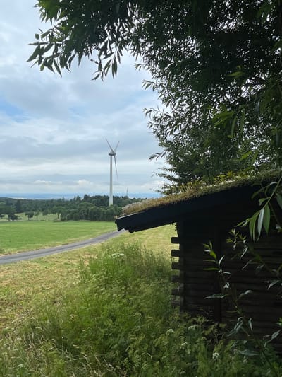 In Schwarzenborn, Germany, a rustic wooden cabin with a moss-covered roof sits in the foreground on the right side of the frame. Its dark, weathered logs are visible, and lush green foliage encroaches upon its roofline and surrounds its base. A paved road winds its way through a verdant, grassy field to the left of the cabin, leading towards a distant landscape of rolling green hills and scattered trees. In the middle ground, a large white wind turbine stands tall against a cloudy sky. The overall impression is one of rural tranquility, with the man-made structures integrated into a natural, picturesque setting.