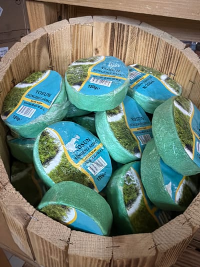 The image shows a close-up, overhead view of a wooden barrel filled with several individually wrapped, round green soaps. The soaps are packaged in plastic with a blue and yellow label featuring an image of moss on a rocky surface and text that reads "YOSUN SÜNGERLI SABUN MOSS SOAP 120ge". The barrel appears to be located in a retail setting, possibly a shop in the city of Acıpayam, Turkey, as evidenced by the text on the packaging. The lighting is natural and appears to be coming from above. The background is blurred but suggests a rustic or naturalistic theme.