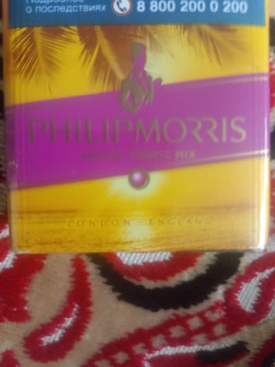 This media file shows a close-up view of a cigarette pack with a tropical theme. The pack is predominantly yellow and purple. Across the top of the pack, a blue band contains the text "о последствиях" and a phone number "8 800 200 0 200". A silhouette of a person is visible against the yellow background. The brand name "PHILIP MORRIS" is prominently displayed in gold lettering on the purple band. Below it, smaller gold text reads "AROMA TROPIC MIX". At the bottom of the pack, the words "LONDON ENGLAND" are visible. The pack appears to be sitting on a patterned fabric, possibly a blanket or rug, with red, black, and white designs. The lighting suggests it is indoors, and there are no clear indicators of time of day or weather. The overall impression is of a product advertisement or packaging.