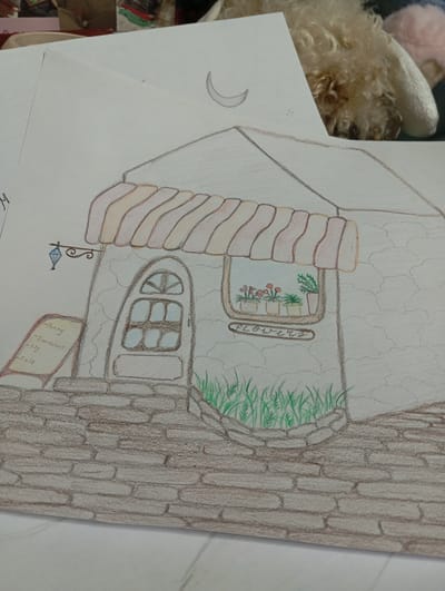 The image is a drawing on paper of a small, quaint shop. The shop has a textured, stone-like exterior. It features a charming arched doorway with a window pane design, and a larger window above which displays potted flowers. An awning with alternating pink and beige stripes extends over the window. A small, decorative sign hangs to the left of the door, with what appear to be flower names written on it: "Peony," "Dandelion," "Lily," and "Rose." Below the window, a sign reads "Flowers" in an oval shape. The shop is situated on a cobbled path, and there is a small patch of green grass in front of the entrance. The background of the drawing shows a light gray, cracked texture, possibly representing a wall or sky. A faint crescent moon is visible in the upper portion of the drawing. In the upper right of the image, part of a fluffy, light-colored dog is visible, suggesting the drawing is on a table or surface with the dog present. The image is taken from a slightly elevated, angled perspective, looking down and across the drawing. There is no indication of the city of Kokand, Uzbekistan, or any elements that would place the drawing in that specific location.
