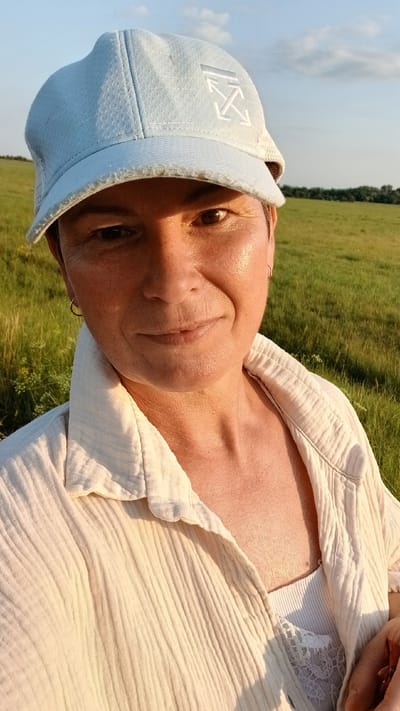 A medium shot, selfie-style photograph features a person wearing a light blue baseball cap and a cream-colored, textured button-up shirt over a white lace-trimmed camisole. The person's face is illuminated by golden hour sunlight, highlighting their features. They are outdoors in a grassy field with a treeline visible in the distance under a partly cloudy sky. The image appears to be taken in the city of Nadezhdenka, Russia.