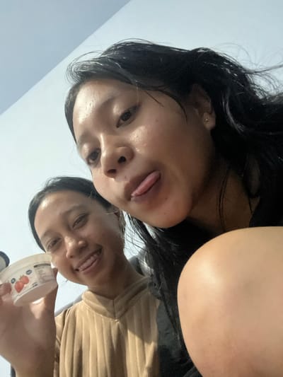 Two young women are captured in a low-angle selfie, appearing to be in an indoor setting, possibly a home, in Jambearum, Indonesia. The woman in the foreground is on the right, her face close to the camera, with her tongue playfully sticking out. Her dark hair is disheveled and appears damp. To her left and slightly behind, another young woman is visible, smiling and holding up a small container of yogurt, which has an image of strawberries on it. She is wearing a tan, ribbed shirt. The background is dominated by a light blue wall. The lighting suggests an ambient indoor environment.