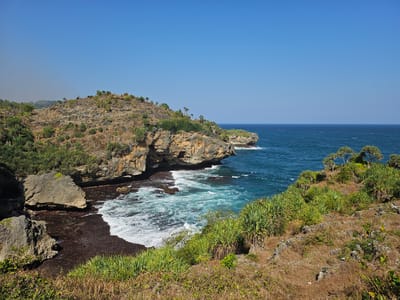 This is a landscape photograph of a rocky coastline in Saptosari, Indonesia, under a clear blue sky. The foreground is dominated by dry, grassy hillsides dotted with low-lying vegetation and some scattered rocks. In the midground, a rugged cliff face juts out into the ocean, with waves crashing against its base and the dark, rocky shore. The water is a vibrant turquoise and blue, with white foam from the breaking waves. In the distance, the ocean stretches to the horizon, with a few small boats visible. The overall impression is one of a wild and beautiful coastal environment.