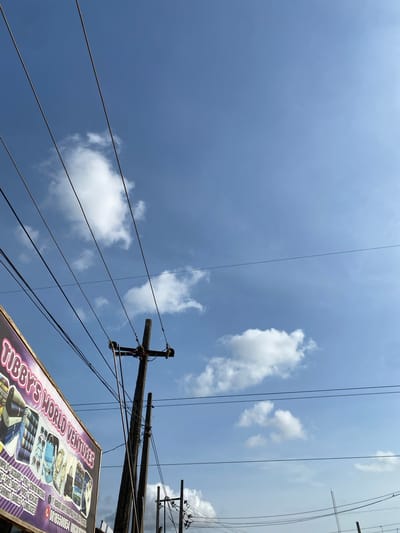 The image depicts a low-angle view toward a bright sky with scattered clouds. Multiple electrical wires run across the frame, connected to wooden utility poles. The lower right corner of the image shows a portion of a sign, identified as "Tubby's World Ventures", which advertises fabrics and accessories. The sign includes a phone number, "08183500541", and an address: "17 Afolabi Jake Road, New B/Stop Ijaiye, Lagos." Thus, the scene is located in the Ijaiye district of Lagos, Nigeria. No people are visible.