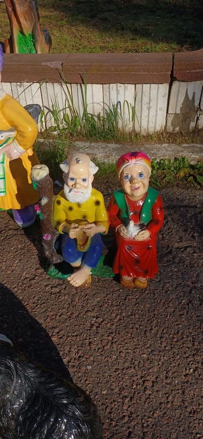 This media file shows a close-up, top-down view of several garden gnome statues placed outdoors. The gnomes are brightly colored and appear to be made of resin or ceramic.
In the foreground and center, an elderly gnome with a white beard wears a yellow shirt and blue pants. He is holding a small basket or container. Next to him, and slightly behind, is a younger gnome with a pink and yellow striped hat and a red shirt with a green collar. This gnome is holding a white bird in its hands. There are parts of other gnomes visible in the lower left and right of the frame, one wearing a yellow shirt and purple pants, and another section of a dark, possibly animal-shaped statue.
The gnomes are positioned on what appears to be a log or textured base, surrounded by dark, gravelly ground. In the background, a section of a white brick wall or pathway is visible, with some green grass and foliage growing nearby. The lighting suggests it is a bright, sunny day, casting distinct shadows. The overall scene evokes a whimsical garden or yard setting. The specific location is Solnechnogorsk, Russia.