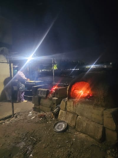 An outdoor oven glows red-orange against the night. Constructed of bricks, the oven sits on a patch of dirt, and a metal plate is visible nearby. A person is seated in front of the oven, illuminated by a bright light source. In the distance, faint lights and trees are seen in the background. The image has a dark and rustic atmosphere, with the oven serving as the central focal point.