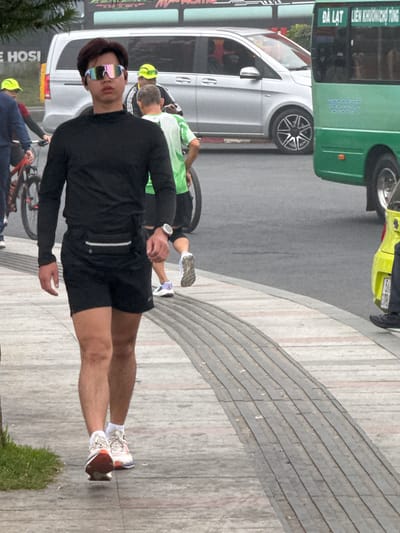 A young man wearing sunglasses, a black long-sleeved shirt, and black shorts walks along a paved sidewalk in Da Lat, Vietnam. He has a fanny pack around his waist and is wearing athletic shoes. Behind him, a silver minivan and a green bus are visible on the street, along with other pedestrians and cyclists. The weather appears to be overcast. The man is walking away from the camera and his face is partially obscured by his sunglasses. The sidewalk has a patterned design of gray and pink tiles.