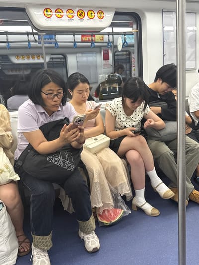 The image shows the interior of a subway car, with multiple individuals occupying the space. A woman with glasses and a white shirt is looking at her cell phone, which she is holding in her hands. Next to her, another woman is reclining on a pillow while using her cell phone. The subway car features warning signs and numerous handrails. In the front of the subway car are a group of younger people who are sitting together. Near one of them there appears to be half of a watermelon.