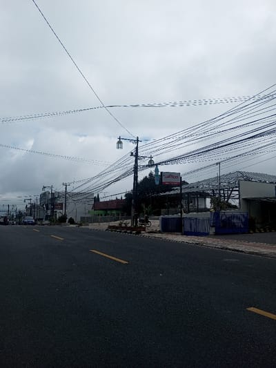 An exterior long shot depicts a daytime street scene in Thotagamuwa, Sri Lanka, under an overcast sky. The asphalt road, marked with yellow dashed lane lines, recedes into the distance from the lower left to the center. On the right side of the road, numerous utility poles support a dense network of overhead black electrical and communication wires that crisscross the upper portion of the frame. A prominent utility pole in the mid-ground features two ornate, cage-lantern style streetlights. Along the right curb, a sidewalk borders a series of structures. Closest to the foreground on the right is a building under construction or renovation, exhibiting exposed metal roof trusses and fronted by a blue and white striped metal fence. Adjacent to this, a sign with "HOTEL YUN" in white text on a red background is mounted on a pole. Further down the street on the right, a dark-colored motorcycle is parked on the sidewalk, followed by a single-story building with a reddish-brown roof, and then a green-colored building. Trees and foliage are visible behind these structures. On the left side of the road, several vehicles, including a blue car, are visible in the distance, alongside multiple buildings and additional utility poles. No individuals are discernible within the frame.