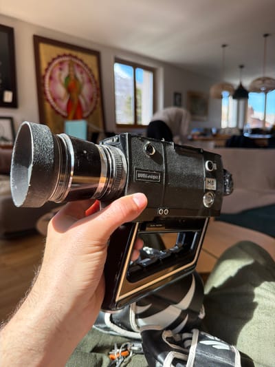 A light-skinned, hairy right hand holds a black Miradge M8 Super 8 film camera in a residence located in Briançon, France. The camera features a textured black lens hood, chrome barrel rings, a black body with "Miradge" and "M8" branding, and a dark brown handle grip with a red trigger button visible under the index finger. A "SLOW MOTION" switch is discernible on the camera's side. Below the camera, portions of olive-green trousers, black and white sneakers, and a set of keys with an orange clip are visible in the foreground. The background, largely out of focus, shows an indoor living space. A large, ornate gold-framed painting depicting a multi-armed Hindu deity within a lotus motif hangs on a light-colored wall to the left. A blurred individual wearing a light-colored top and dark lower garments is bent over in the mid-ground. Natural light streams into the room through at least two windows, revealing a clear blue sky outside and casting distinct shadows within. Two woven pendant light fixtures hang from the ceiling, and indistinct furniture, including a couch, is present further in the background.