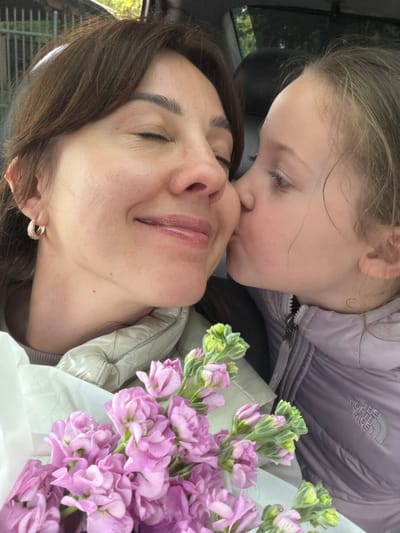 A young girl with light hair is kissing a woman with brown hair, who is lying down with her eyes closed. The girl is wearing a lavender-colored jacket with "The North Face" logo on the chest. Next to them is a bouquet of purple and green flowers. The picture appears to be taken inside a car. Location cannot be determined from the image, so the query about Salaspils, Latvia cannot be answered.