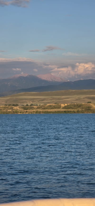 The image depicts a wide view of a body of water in the foreground, with a textured surface indicative of ripples. Beyond the water, a gently sloping, treeless landscape stretches towards a range of mountains in the distance. The mountains exhibit varied geological formations and are partially obscured by clouds. The sky above is a pale blue, with scattered, soft clouds that are illuminated by a warm light, suggesting either sunrise or sunset. There are no people or discernible man-made structures in the immediate vicinity of the water, nor in the middle ground landscape. The scene evokes a natural, serene environment. The location is not identifiable as Pristan'-Przheval'sk, Kyrgyzstan, based on the visual evidence presented in the image.