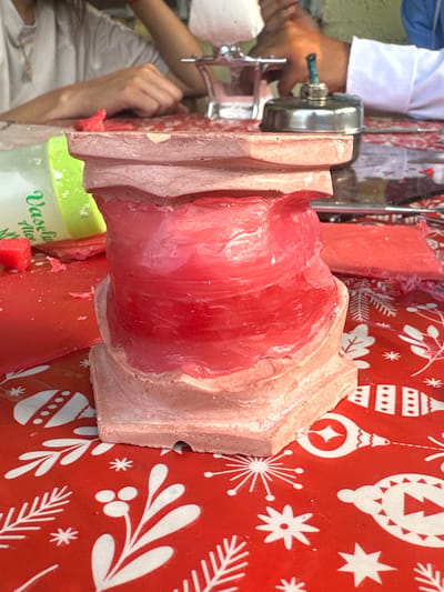 The image shows a process of making wax candles. A block of pink wax is being compressed through a shaping device, likely a manual press, which has a silver handle. There is a person in the background operating the press, wearing a white sleeve. A jar of Vaseline Aloe Fresh is sitting near the wax block. The entire scene is set on a red surface printed with white Christmas-themed designs such as ornaments, snowflakes, and trees. The setting appears to be indoors, possibly a workshop or studio. This is in Parroquia Ocumare, Venezuela.