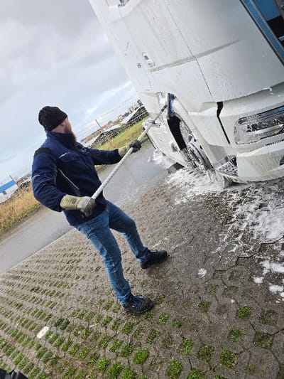 In Ostend, Belgium, a bearded male is depicted actively washing a white semi-truck. The individual wears a black beanie, a dark blue work jacket with lighter accents, blue jeans, dark shoes, and light-colored work gloves. He utilizes a long-handled white and blue brush to scrub the lower front passenger side of the truck's cab, which is extensively covered in white cleaning foam. Water and soap suds splash onto the wet ground, which consists of concrete grid pavers with interspersed grass, alongside areas of wet asphalt. The outdoor scene is under an overcast sky, with a wire fence, a grassy field, and distant solar panels visible in the background.