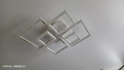 A modern, multi-tiered square ceiling light fixture is mounted on a plain white ceiling. The fixture is composed of several interlocking squares of varying sizes, outlined with small, reflective embellishments that appear to be crystals or rhinestones. The light fixture is white and appears to be made of metal or a similar rigid material. The overall impression is one of contemporary design. The image is watermarked in the lower left corner with "Infinix ZERO 30". The location of Bugulma, Russia is not discernible from the visual information provided in the image.