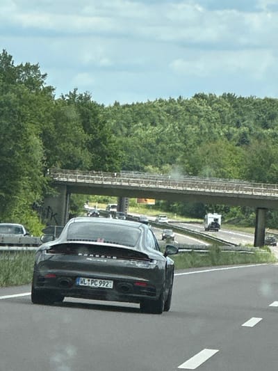 The image depicts the rear of a black Porsche 911 (992) driving on a highway. The license plate is visible and reads "WL PC 992". A highway overpass is visible, with trees and greenery in the background. The sky is partly cloudy. Based on the license plate and general environment, the location is consistent with a highway in the general vicinity of Buchholz in der Nordheide, Germany.