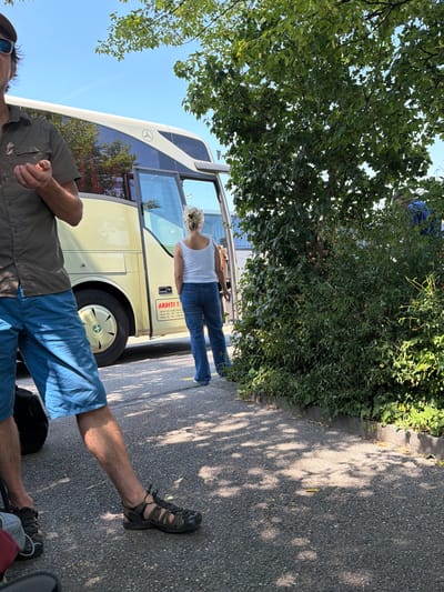 This image shows a sunny outdoor scene with two people and a bus. The primary focus is on a person in the foreground, partially visible, wearing blue shorts and a grey shirt. Their legs and feet are prominent, with one leg extended and wearing an open-toed sandal. Behind them, a woman stands by an open door of a light-colored bus, wearing a white tank top and blue jeans. The bus has a Mercedes-Benz logo on its side.

The background features green foliage and trees, suggesting a natural or park-like setting. The ground is paved with asphalt, and dappled sunlight creates shadows on the surface, indicating a bright, sunny day. The overall atmosphere appears relaxed and casual.

Visible text on the bus reads "ARDITI T" followed by contact information including mobile numbers for Kosovo, Switzerland, and Albania. The presence of these numbers and the Mercedes-Benz logo suggest the bus is likely a tour or transportation vehicle. The location context is Lausanne, Switzerland, which aligns with the presence of a Swiss phone number. The time of day appears to be daytime, likely late morning or afternoon, given the strong sunlight and shadows. No specific emotions are explicitly conveyed by the people, but the scene suggests a moment of transition or waiting.