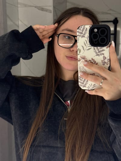 A female is depicted in a mirror selfie in Prikubanskii, Russia, captured from the chest up. She has long, straight brown hair, wears black-framed clear glasses, and is dressed in a dark gray, faded zip-up hooded sweatshirt over a dark undershirt. Her left hand is raised to her temple. Her right hand holds a smartphone, partially obscuring the lower right portion of her face. The phone, an iPhone model with a prominent triple-lens camera array, is encased in a cream-colored cover patterned with brown and green botanical motifs and butterflies. A metallic ring with small embedded stones is worn on her right ring finger. The background consists of light-colored, rectangular wall tiles with subtle veining, and a dark vertical fixture is partially visible in the upper right.