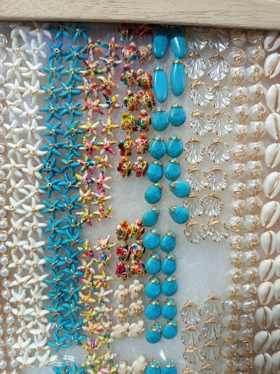 The image displays a collection of assorted jewelry components, predominantly arranged in vertical columns within a display case. The components include white pearl-like beads, blue starfish-shaped charms, colorful, multi-hued starfish charms, small turtle-shaped charms with colorful designs, teardrop-shaped turquoise stones, clear shell-shaped charms with gold accents, and white cowrie shells with gold accents. The display case is framed by a light-colored wooden border at the top. The overall impression is of a retail display of craft or jewelry-making supplies. The scene is not specific to Tolú, Colombia, and no people, actions, or interactions are depicted.