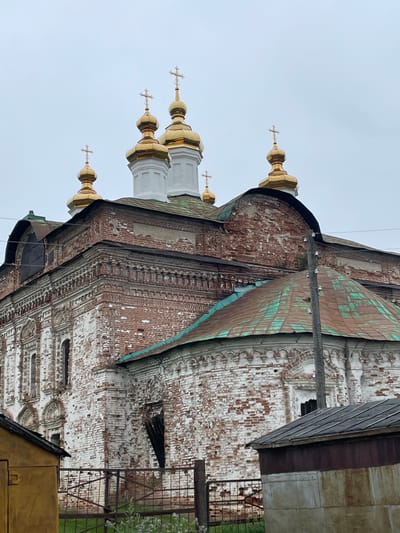 The image shows a close-up, low-angle view of the brick facade of an old, Orthodox church, likely located in Kstynino, Russia. The church is weathered, with areas of red brick exposed through peeling white paint. Several golden domes with crosses are visible at the top of the structure, indicating its religious purpose. One of the domes has a green, oxidized patina, suggesting age. The church has ornate architectural details, including decorative brickwork and arched window frames, many of which are dark and appear to be boarded up or empty. In the foreground, parts of simpler, more modern structures, including a yellow building and a corrugated metal shed, are visible. A metal fence runs across the lower part of the image. A utility pole with insulators stands to the right of the church. The sky is overcast, suggesting a cloudy day. No people are visible in the image.