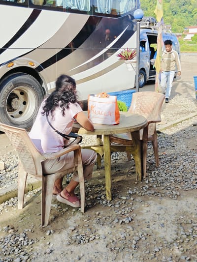 In Tharpu, Nepal, a woman sits at a round plastic table on a gravel-covered ground, facing away from the camera. She has curly dark hair and wears a light pink t-shirt and light-colored pants. On the table, an orange and white reusable shopping bag with the word "MALLA" is visible. She is seated in a beige plastic chair. Another identical chair is placed next to the table. Behind the table, a large bus with a black and silver design is parked, its rear tire prominent in the frame. To the right of the bus, a man in a light brown hooded sweatshirt and light blue jeans walks away from the camera. A small blue van is parked behind the bus, and colorful flags hang on a pole between the bus and the van. The environment appears to be an outdoor waiting area or rest stop, with a mountainous backdrop in the distance.