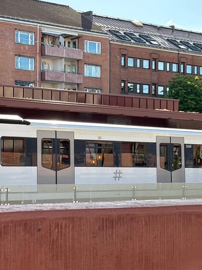 An outdoor, medium shot captures a train car with its doors and windows visible, positioned in front of a multi-story brick building with balconies and a mansard roof. The train car is white with dark gray sections around the windows and doors. A number, "3260", is visible on the right side of the train car, along with a hashtag symbol on the left side. The building features rows of windows and balconies, some of which appear to be in disrepair. The sky above is a clear, light blue. In the foreground, a reddish-brown railing runs horizontally across the image.