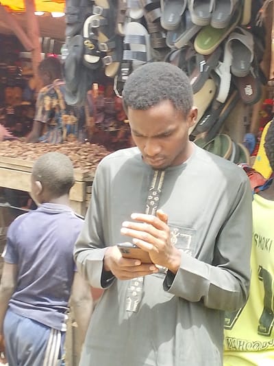 The image captures an outdoor market scene in Fagge D1, Nigeria. A dark-complexioned adult male, centrally positioned, wears a gray traditional long-sleeved garment with white and gold embroidery. He is focused on a light brown smartphone held horizontally in both hands. To his left and slightly behind, a younger individual, seen from the back, wears a dark blue short-sleeved polo shirt and blue shorts with white stripes. Behind them, a wooden stall displays numerous small, brown, oval-shaped items, identified as nuts or seeds, with a vendor in a patterned shirt partially visible behind it under an orange awning. To the immediate right of the central figure, a person in a bright yellow shirt with black lettering ("SHOKI") is partially visible. Above and behind the central figure, a vertical display rack is laden with various pairs of slippers and sandals, indicating a footwear stall.