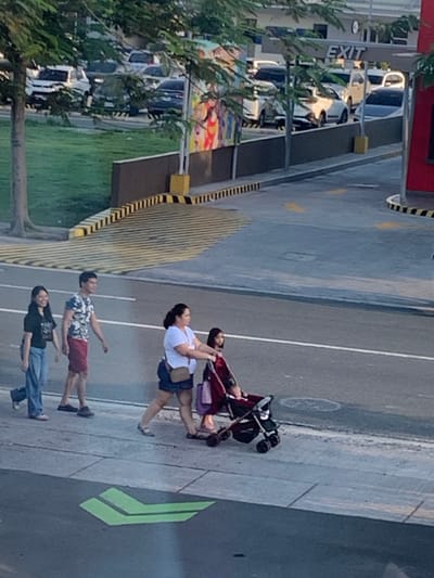 In Angeles City, Philippines, a low-angle medium shot captures a sidewalk and road scene. On the sidewalk, four individuals are visible. From left to right, a young woman in a black t-shirt and blue jeans walks with a slight smile. Next to her, a man in a camouflage t-shirt and red shorts walks, also with a subtle smile. Further to the right, a woman wearing a white t-shirt and dark shorts pushes a stroller containing a young child dressed in red. The stroller is dark-colored with red accents. Behind them, another child is partially visible in the stroller. The group appears to be walking together on the sidewalk adjacent to a paved road. In the background, cars are parked in a lot, and an "EXIT" sign is prominent. Trees and greenery are scattered throughout the scene, and a brightly colored mural is visible on a building in the distance. Yellow and black striped barriers are present along the edge of the road and sidewalk. A green arrow painted on the road indicates a direction. The overall lighting suggests it is daytime.