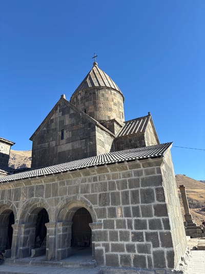 A stone building, possibly a church or monastery, features a dome topped with a cross against a clear blue sky. The structure showcases detailed stone masonry, including arched windows and a roof with a textured edge. A section of the building presents a facade of tightly fitted stone blocks. A mountainous landscape is visible in the background, enhancing the building's architectural details.