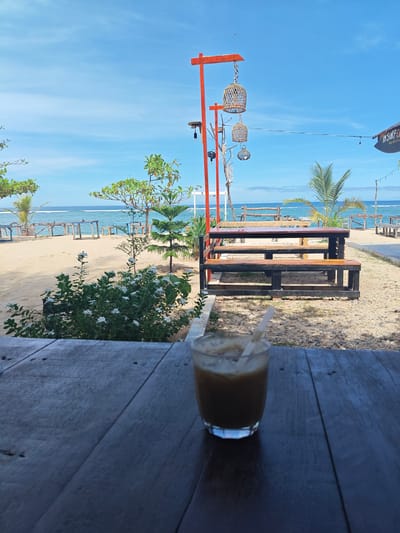 The image shows a beachside scene in Monikeun, Indonesia. A glass of iced coffee with a straw is placed on a dark wooden table in the foreground. Beyond the table, the scene opens up to a sandy area with several wooden picnic tables and benches. Green trees and bushes are interspersed throughout the sand. The ocean is visible in the distance, with small waves and a clear blue sky above. There is a tall orange structure with hanging wicker decorations. A sign reading "IM SURF CAFE" is partially visible on the right side of the frame.