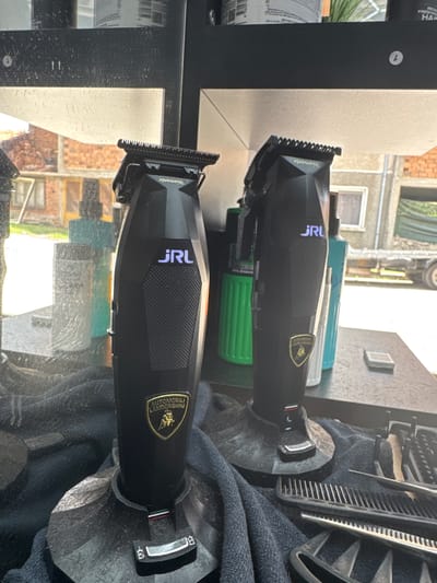 This is a close-up, eye-level shot of two black JRL professional hair clippers with Lamborghini logos, placed on black stands. The clippers are positioned on a dark countertop, with a dark blue towel beneath and to the right. Various barber tools, including combs and scissors, are visible on the towel to the right of the clippers. In the background, out of focus, are shelves with bottles of hair products and a window looking out onto buildings constructed of red brick, suggesting an outdoor scene in the city of Dobrinishte, Bulgaria. The lighting is natural and bright, indicating it is daytime. There are no people present in the image.