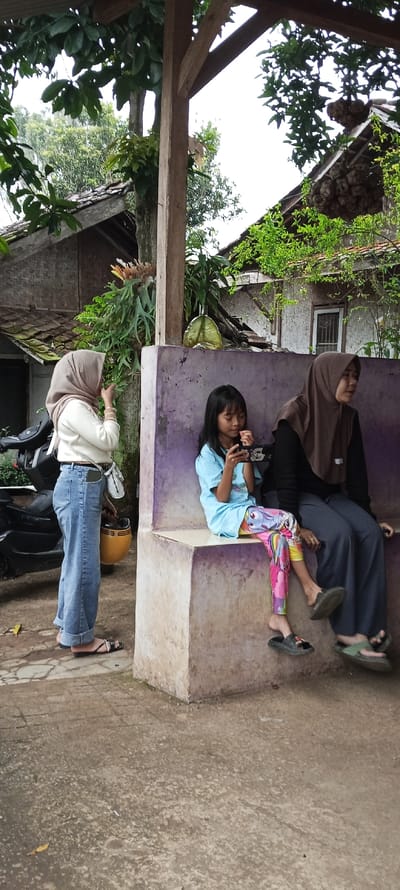 Three women are standing in front of a concrete bench. The bench is situated under a wooden pergola with a partially obscured house behind it. The woman standing closest to the camera is wearing blue jeans, a white long-sleeved shirt, and a headscarf. She is holding a yellow helmet and appears to be talking to the other woman sitting on the bench. The woman on the bench is wearing a long-sleeved blue shirt and colorful pants. She is holding a small electronic device.  The third woman is seated on the bench wearing a brown headscarf and a black long-sleeved shirt. She is looking directly at the woman standing to her left. The scene is in Cimaung, Indonesia, with coordinates -7.074 latitude and 107.547 longitude.