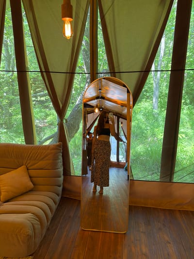 A woman stands in a tent-like structure and takes a mirror selfie. She is wearing a black tube top and a leopard print skirt. The tent has large windows with green fabric curtains, revealing a dense forest outside. A large, tufted sofa is visible on the left side of the frame. The floor is made of dark wood planks. A gold-framed mirror is positioned in the center of the frame. The scene appears to be a glamping tent in a forest setting.