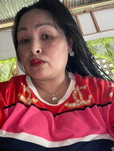 A woman with dark hair, red lipstick, and jewelry is in Hijo, Philippines. She wears a top with red, pink, black, white, and yellow patterns. The background includes what appears to be a window with a green and white curtain or covering.