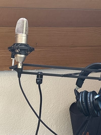 The image displays a static audio recording setup located in Hagen, Germany. A silver-toned condenser microphone, encased within a black circular shock mount, is positioned in the upper left quadrant. This microphone assembly is secured to a black articulated boom arm, which extends horizontally across the frame and incorporates visible tension springs. The boom arm is clamped to an unseen horizontal surface, likely a desk or shelf edge. A black XLR cable connects to the base of the microphone and descends downwards. To the right, a pair of black over-ear headphones is suspended from a component of the boom arm. The background consists of two distinct horizontal elements: a light-colored, textured wall forming the lower section, and horizontal light-brown wooden paneling or a wooden surface comprising the upper section. No individuals are present, and no active actions are depicted.