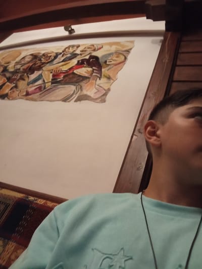 A young person, possibly a male, is shown from the chest up, wearing a light blue crewneck sweatshirt. They have dark hair and are looking to the right, with their ear visible. In the background, a painting depicting a group of people in a stylized, somewhat abstract manner is mounted on a light-colored wall. The painting appears to be in a warm color palette with earthy tones and some red and blue accents. The overall environment suggests an indoor setting, possibly a restaurant or a home, with wooden beams and trim visible at the top and right of the frame, and a patterned fabric, perhaps upholstery or a blanket, in the lower left corner. The image does not provide specific details to pinpoint the location within the city of Devin, Bulgaria, beyond the implied interior setting.